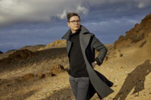 Pianist Vikingur Olagsson wears a grey coat as he stands outdoor on rocky, sandy terrain with a blue sky visible above him.