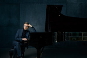 Pianist Vikingur Olaffson sits against a black grand piano and props up his head on his hand, wearing a blue suit.