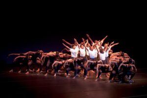 A performance in Aileycamp with a large dance ensemble, the dancers in the center wearing white shirts and strikingly extending their arms.