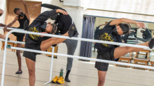 Dancers from Aileycampt stretch their legs along a ballet barre.