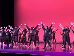 Aileycamp members wear the Aileycamp merch on a stage lit up with bright pink lights, and raise their arms in a final pose.