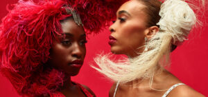 Two dancers from the Alvin Ailey American Dance company pose close to each other with elaborate red and white feather headpieces in their hair.