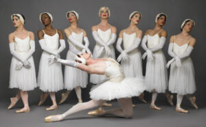 A ballet dancer in a white tutu performing a pose in the foreground with six other dancers in matching attire standing in profile in the background.