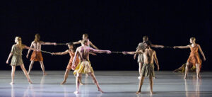 The Mark Morris Dance Group performs in different colored costumes on stage against a beige background.
