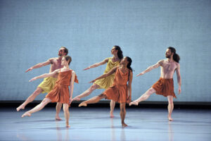 The Mark Morris Dance Group performs in different colored costumes on stage against a beige background.