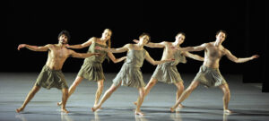 Mark Morris Dancers wear grey costumes and dance in a line formation on stage.