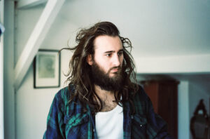 Jean Rondeau pictured in his home, wearing a flannel shirt with long hair and a beard.
