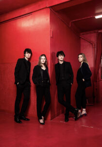 The four members of the Le Consort Courtesy group wear black formal dress, standing in a bright red room.