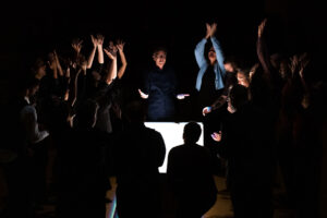 Members of the Los Angeles Master Chorale stand around a lighted box in a dark room.