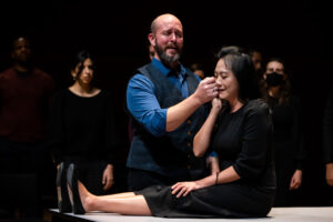 Two performers embrace on stage, a man caresses a woman's cheek as she sits down next to him.
