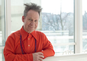 Peter Sellars wears a bright orange shirt with his hair standing straight upwards.