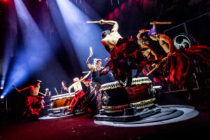 Drummers leap in the air while beating large drums on stage in the Drum Tao 30th anniversary performance, wearing vibrant red costume skirts.