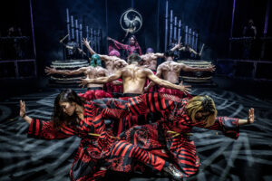 Members of the Drum Tao Group pose in front of each other on stage, leading to the large drums in the back of the group. The female members in front wear vibrant red and black striped robes and the male members in the middle stand with their bare backs to the front and arms raised.