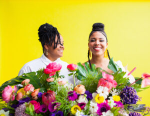Two women in the Okan group smile while holding flowers in front of a yellow background.