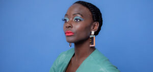 A photo of Nathalie Joachim, wearing her hair in a braided updo, light blue eyeshadow, and geometric rectangle earrings.