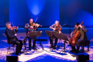 The Kronos Quartet in a live performance, playing instruments on a stage illuminated by blue and purple neon lights.