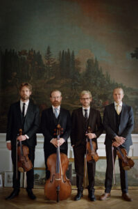 The Danish String Quartet stand together for a group photo, wearing black dress suits and holding their instruments in front of a large painting of a forest.