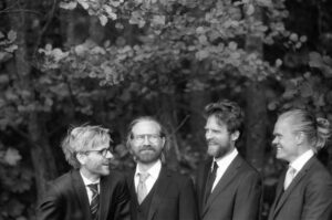 The Danish String Quartet stand together in this black and white photo amidst a lush forest background of trees.