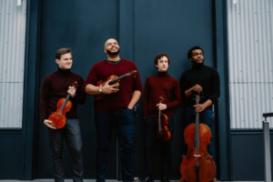 The Isidore String quartet stands with their instruments in front of a blue set of doors.