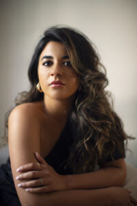 Amina Edris sits in this photo with her voluminous hair in waves, and wears a one-shouldered top.