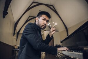 Conrad Tao sits and plays piano with one hand, the other raised in a gesture.