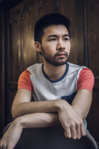 A close up image of artist Conrad Tao with his arms folded in his lap, wearing casual clothing and looking off camera.