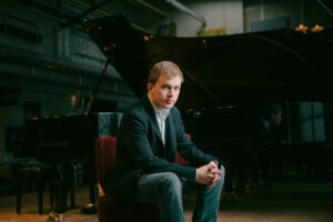 Artist Fillipo Gorini wears a black suit and sits on a red seat in front of a grand piano