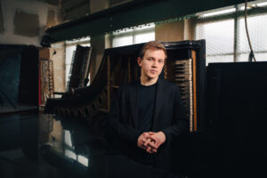 Artist Fillipo Gorini wears a black turtleneck and suit jacket, standing in front of an art installation of a piano turned on it's side.