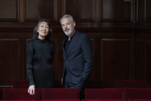 Mitsuko Uchida and Mark Padmore stand next to each other wearing navy blue formal wear in front of velvet red theater seats.