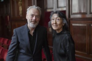 Mark Padmore and Mitsuko Uchida stand together in a wooden room.