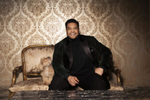 Pene Pati sits in an ornately decorated room, on a small couch with a tan dog sitting next to him.