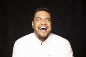 A photo of singer Pene Pati wearing a white shirt against a black background, laughing animatedly.