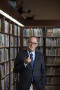 David Sedaris wearing a navy blue suit and blue patterned tie, standing with a pipe in his hand in front a corner of tall bookshelves.