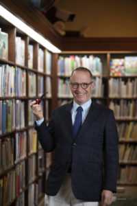 David Sedaris wearing a navy blue suit and blue patterned tie, smiling largely with a pipe in his hand in front a corner of tall bookshelves.