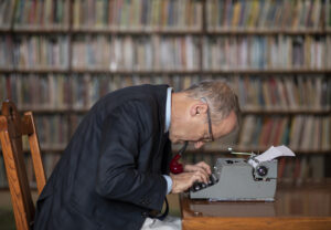 David Sedaris sitting with a pipe in his mouth, leaning over a typewriter at a desk in front of a wall of booskelves.