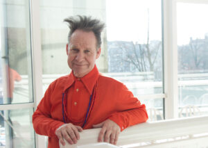 Peter Sellars wears a bright orange shirt with his hair standin straight upwards.
