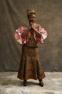 Angelique Kidjo stands in a beige room wearing an elaborate multicolored dress and matching headwrap.