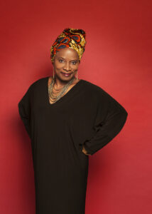 Angelique Kidjo wears a flowy black dress and multicolored yellow and red headwrap while standing against a bright red wall.