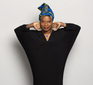 Angelique Kidjo wears a flowy black dress and holds both her arms up to her head next to her blue and yellow headwrap.