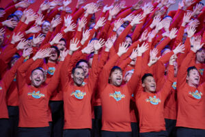 The men of the San Francisco Gay Men's Chorus stand on risers and sing while all raising their arms above their head and wearing matching red shirts.