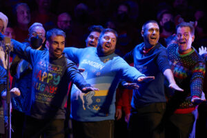 Members of the San Francisco Gay Men’s Chorus wear Holiday sweaters and sing together with their arms extended outwards.
