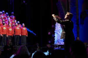 The conductor stands in front of the San Francisco Gay Men's Chorus in red sweaters.