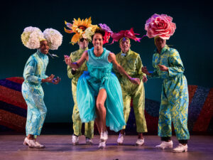 Performers in the Dorrance Dance group wear large flower headpieces and configure together in a tap-dance routine on stage.