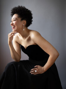 Artist Julia Bullock wears an elegant strapless dress and pearl jewelry, and her side profile shows her smiling brightly off screen.