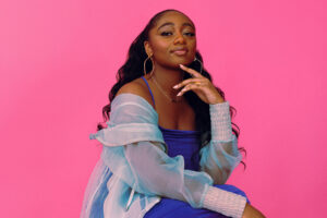 Jazz singer Samara Joy, a young black woman with long wavy hair, wears a bright blue dress and overshirt that contrasts vividly with the hot pink background.