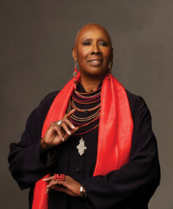 Judith Jamison, a black woman with a shaved head wearing a bright red neck scarf with jewelry that match in color, looking into the camera