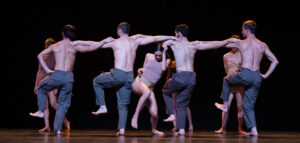 Shirtless male dancers from Batsheva Dance Company link arms during a performance.