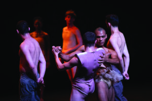 Members of the Batsheva dance company, a combination of shirtless or in nude body suits, dance around two members engaged in a pair dance.