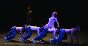 Members of the Batsheva dance troupe onstage crawling on all fours in a line with two dancers standing behind them.