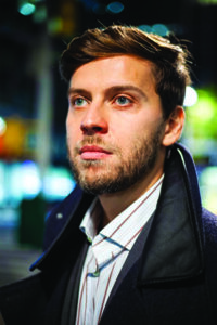 Vladimir Rumyantsev, a Russian man with short brown hair wearing a striped button-up and black coat, looks off to the side of the camera.